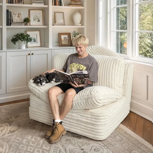 Młoda osoba siedząca wygodnie w dużym, beżowym fotelu modułowym, sofie rozkładanej (welur prążkowany) i czytająca książkę.