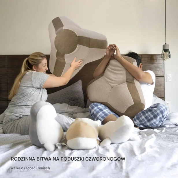 Para dorosłych, siedząc w łóżku, bawi się, prowadząc "bitwę na poduszki" za pomocą dużych, geometrycznych poduch.