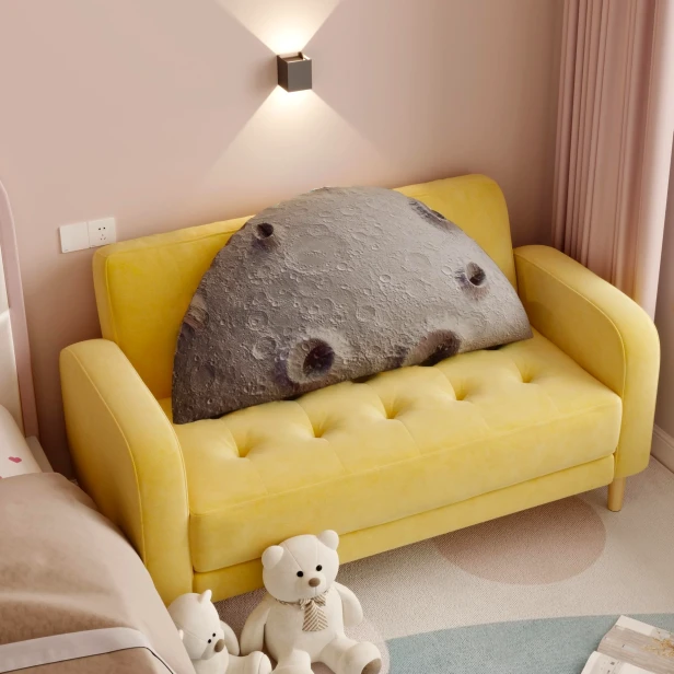 Duża, półokrągła poduszka w szarościach z motywem Księżyca (kratery) 3D leżąca na małej, pikowanej, żółtej kanapie.