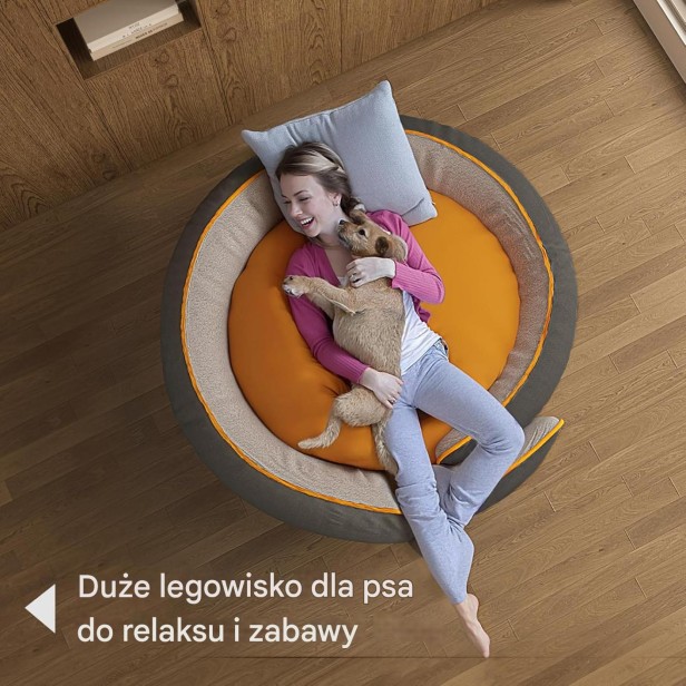 Zdjęcie z góry ukazujące kobietę przytulającą małego psa w bardzo dużym, okrągłym legowisku w kolorze szaro-pomarańczowym.