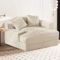Duży, głęboki szezlong, leżanka w beżowym/kremowym kolorze, tapicerowany grubym, miękkim sztruksem z widocznymi prążkami. Mebel posiada dwie dekoracyjne poduszki o tym samym prążkowanym wzorze, ustawiony w eleganckim salonie.