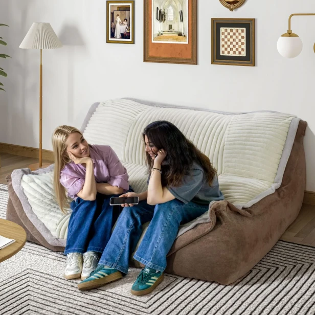 Dwie młode kobiety siedzące na dużej modułowej Kanapie Wypoczynkowej w kolorze brązowym z beżowym, pikowanym pokrowcem.