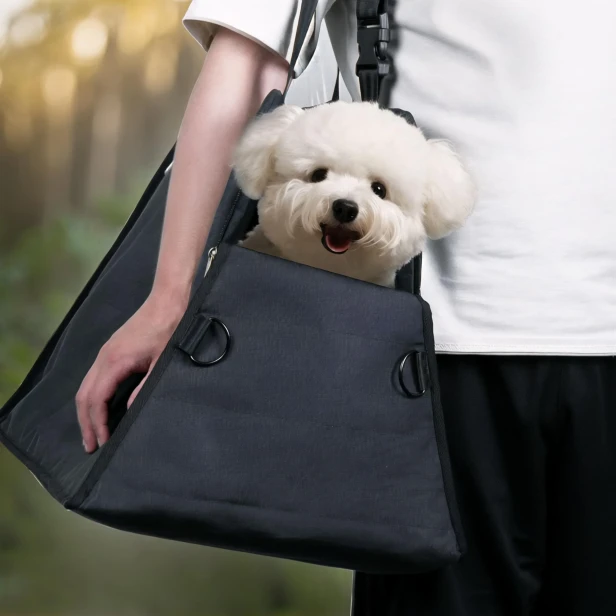Biały pies (Bichon) wystający z czarnej torby-nosidła na ramię, niesionej przez właściciela. Fotelik Podróżny 3w1